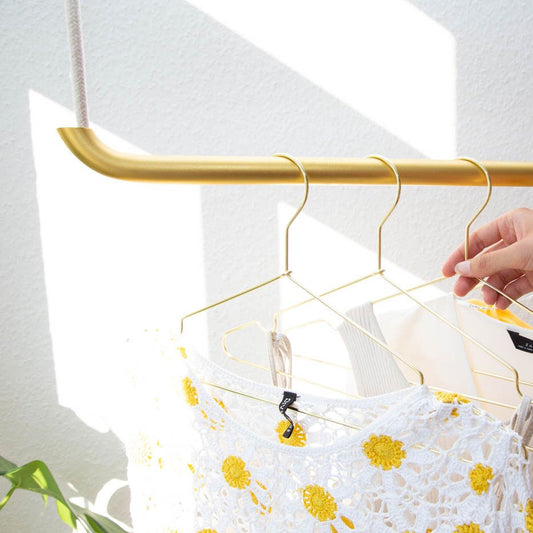 Eine Hand hängt einen goldenen Kleiderbügel mit einem weiß-gelben Häkeldeckchen an die Stab & Knoten Kleiderstange zur Deckenmontage (Metall, 70/110 cm); Sonnenlicht und Schatten spielen auf der weißen Wand dahinter.