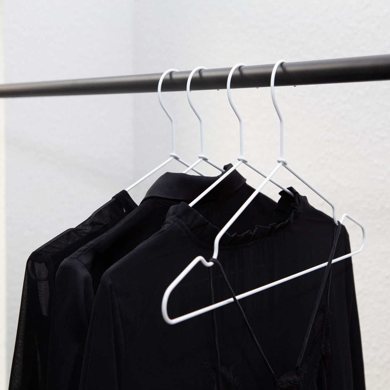 Vier schwarze Kleidungsstücke auf weißen Kleiderbügeln aus stabilem Metall (8 Stück) von Rod & Knot hängen an einer schwarzen Kleiderstange. Die Kleidungsstücke scheinen leicht und durchsichtig zu sein, wahrscheinlich Blusen oder Hemden, die vor einer schlichten weißen Wand ausgestellt sind. Die Kleiderbügel sind gleichmäßig verteilt, was für ein ordentliches und organisiertes Erscheinungsbild sorgt.